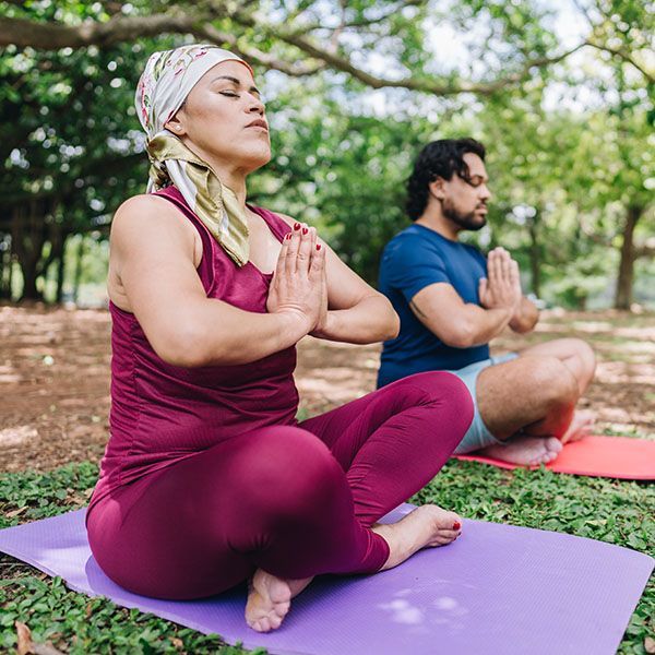 Two people sit cross-legged on yoga mats outdoors, meditating with hands together in a park.