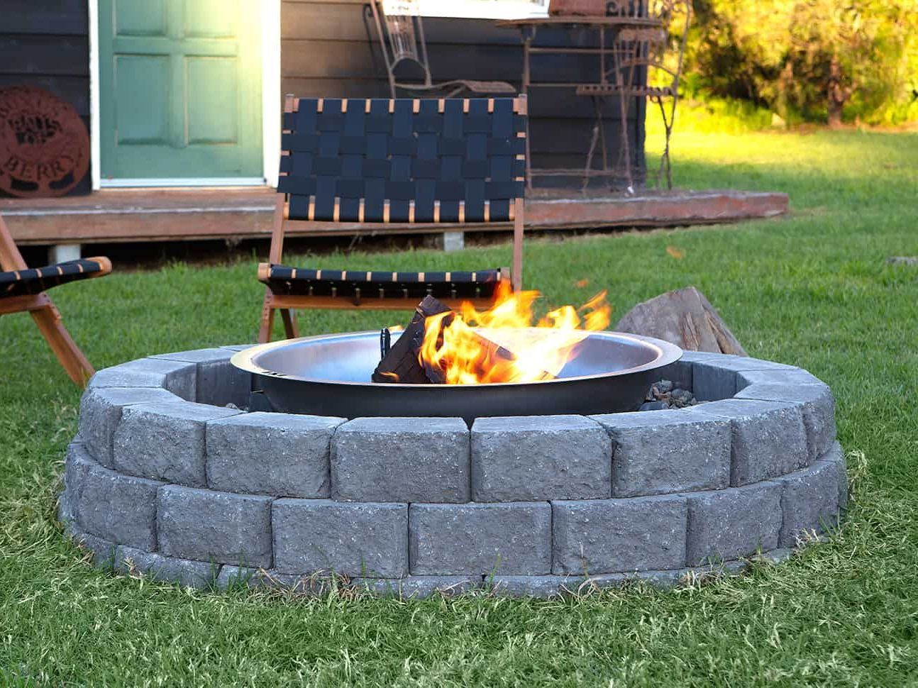 Fire pit with burning flames, built of gray blocks, in a grassy yard, with a chair in background.