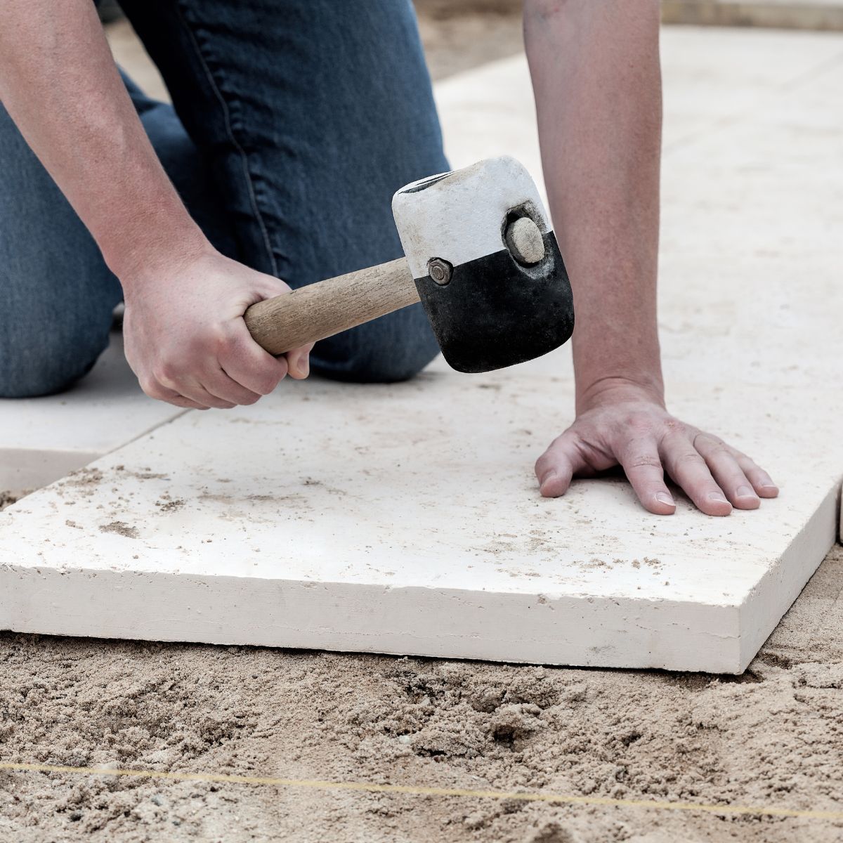 Person uses a mallet to level a light-colored paving stone on a sandy surface.