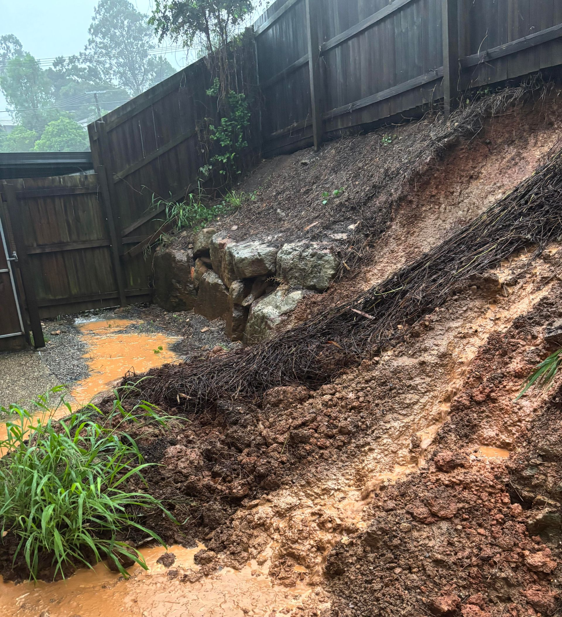 Backyard slope eroded by runoff, with muddy soil, a wooden fence, and standing water.