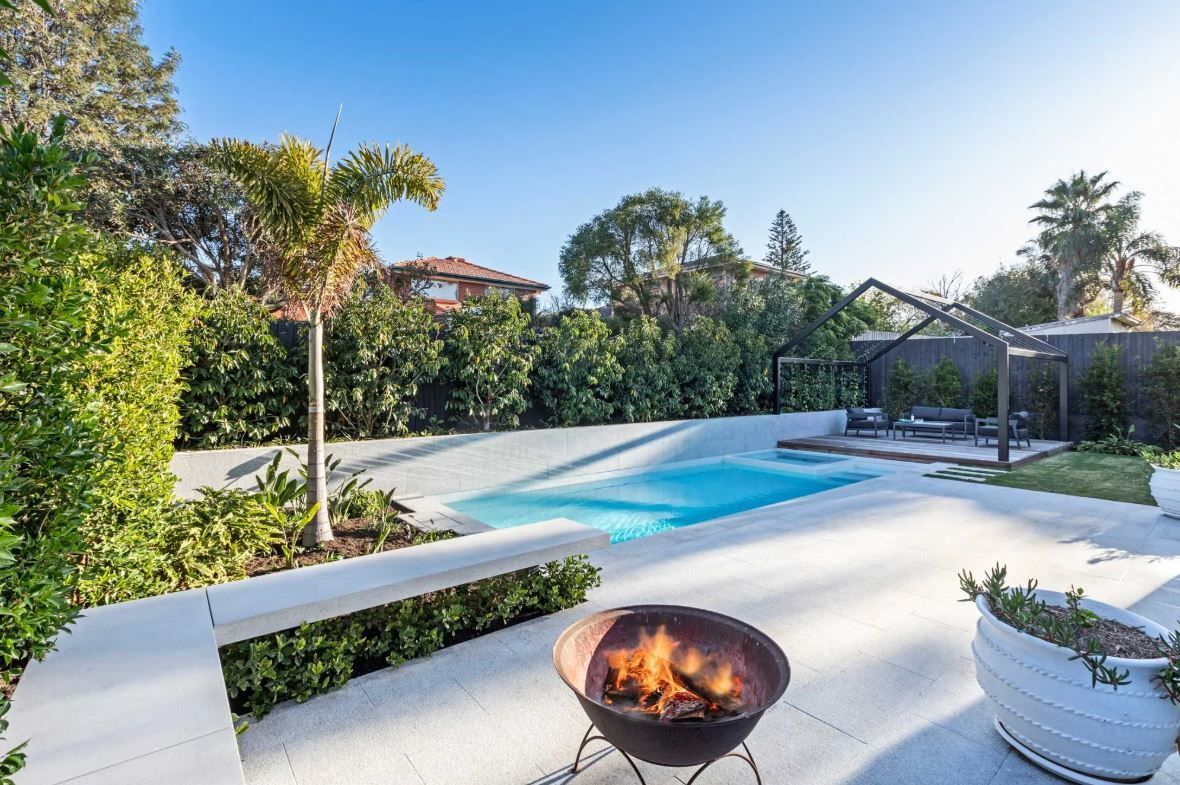 Backyard with pool, fire pit, and lush greenery on a sunny day.
