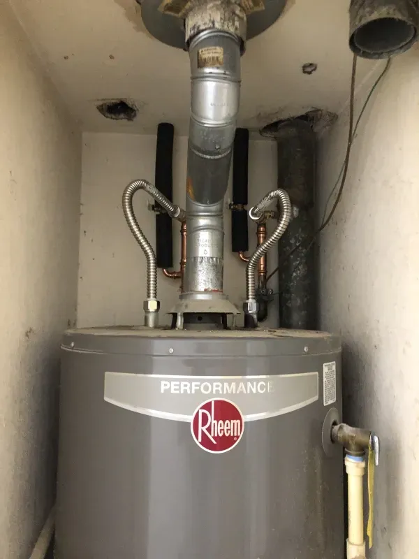 Gray Rheem Performance water heater in a utility closet with exposed pipes and ventilation duct.