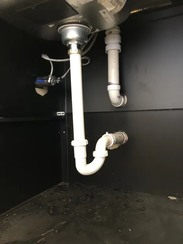 Under-sink plumbing: white pipes, including a P-trap, connected to a stainless steel sink drain. Black cabinet.