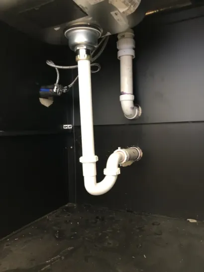 Under-sink plumbing: white pipes, including a P-trap, connected to a stainless steel sink drain. Black cabinet.