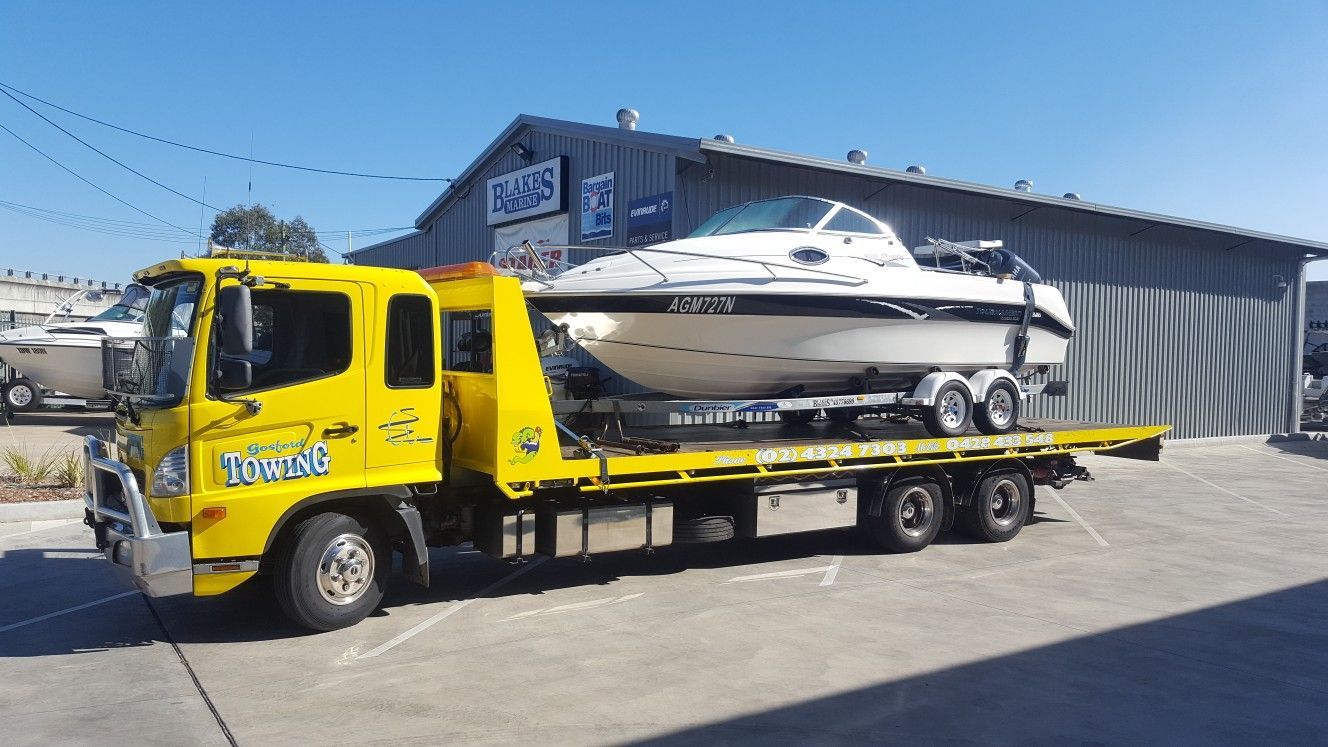 A yellow tow truck is carrying a boat on the back of it.