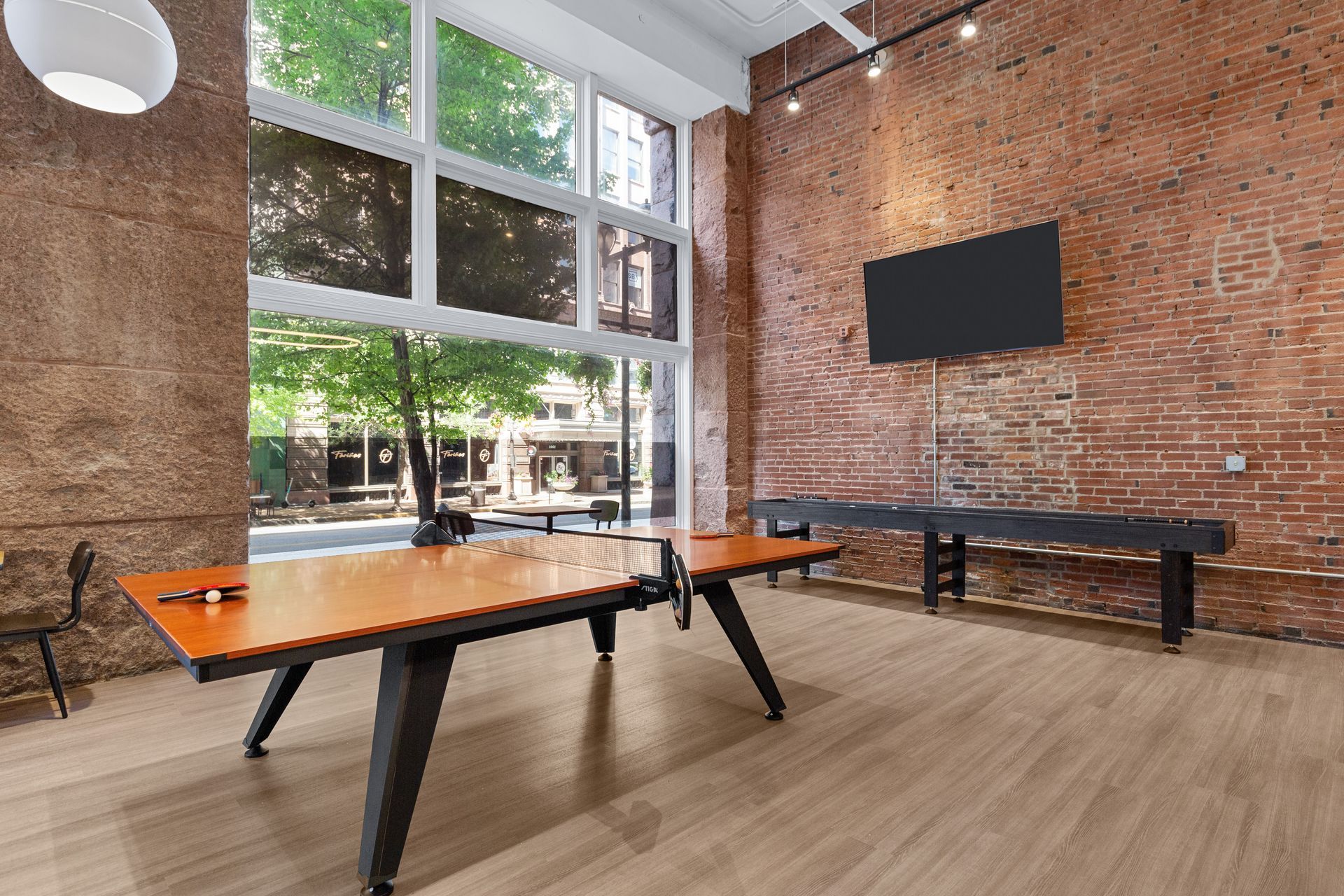 Empty modern room with brick walls, large windows, a TV, and black ping-pong tables on wood flooring