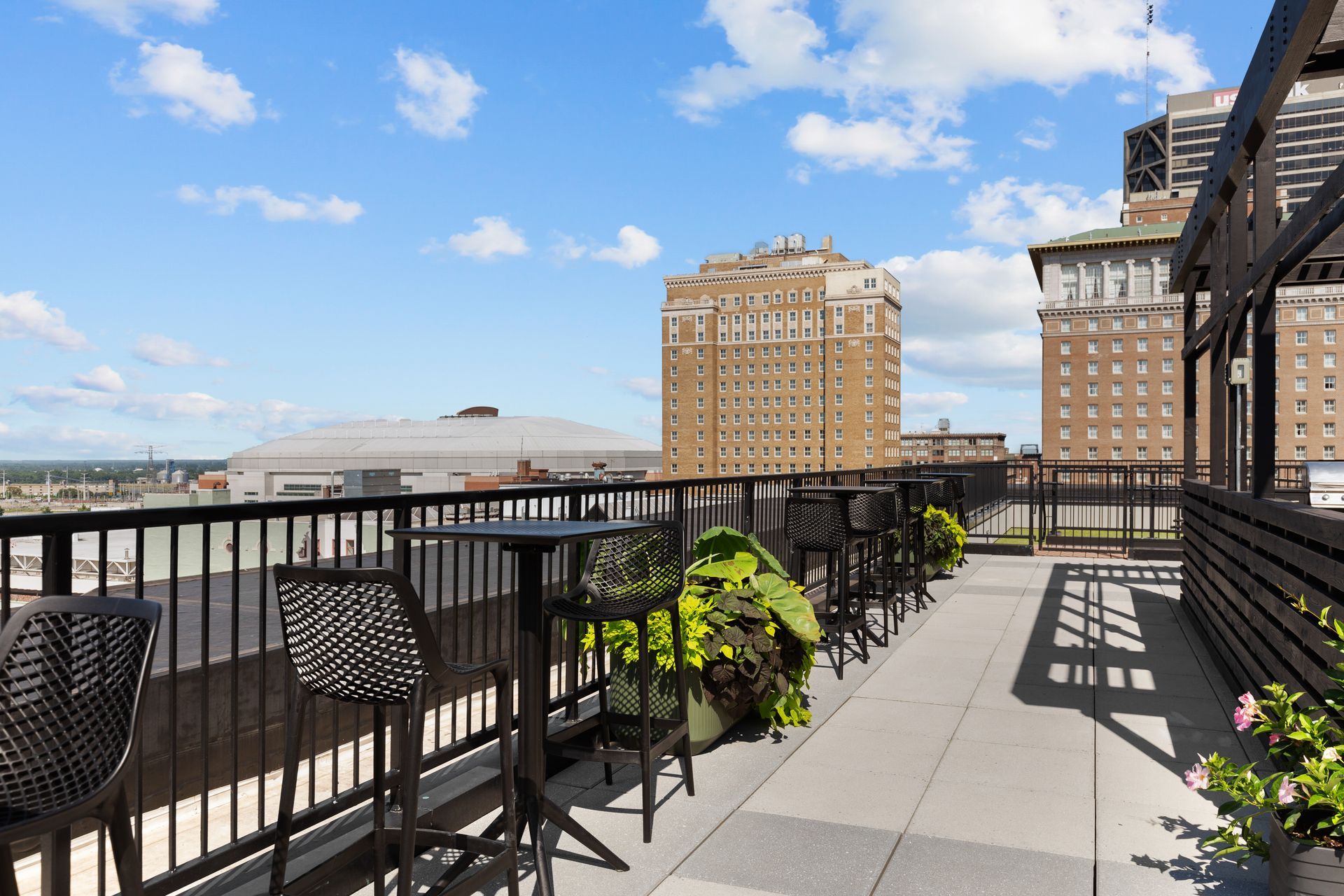 Rooftop terrace with black railings, potted plants, and city buildings under a blue sky