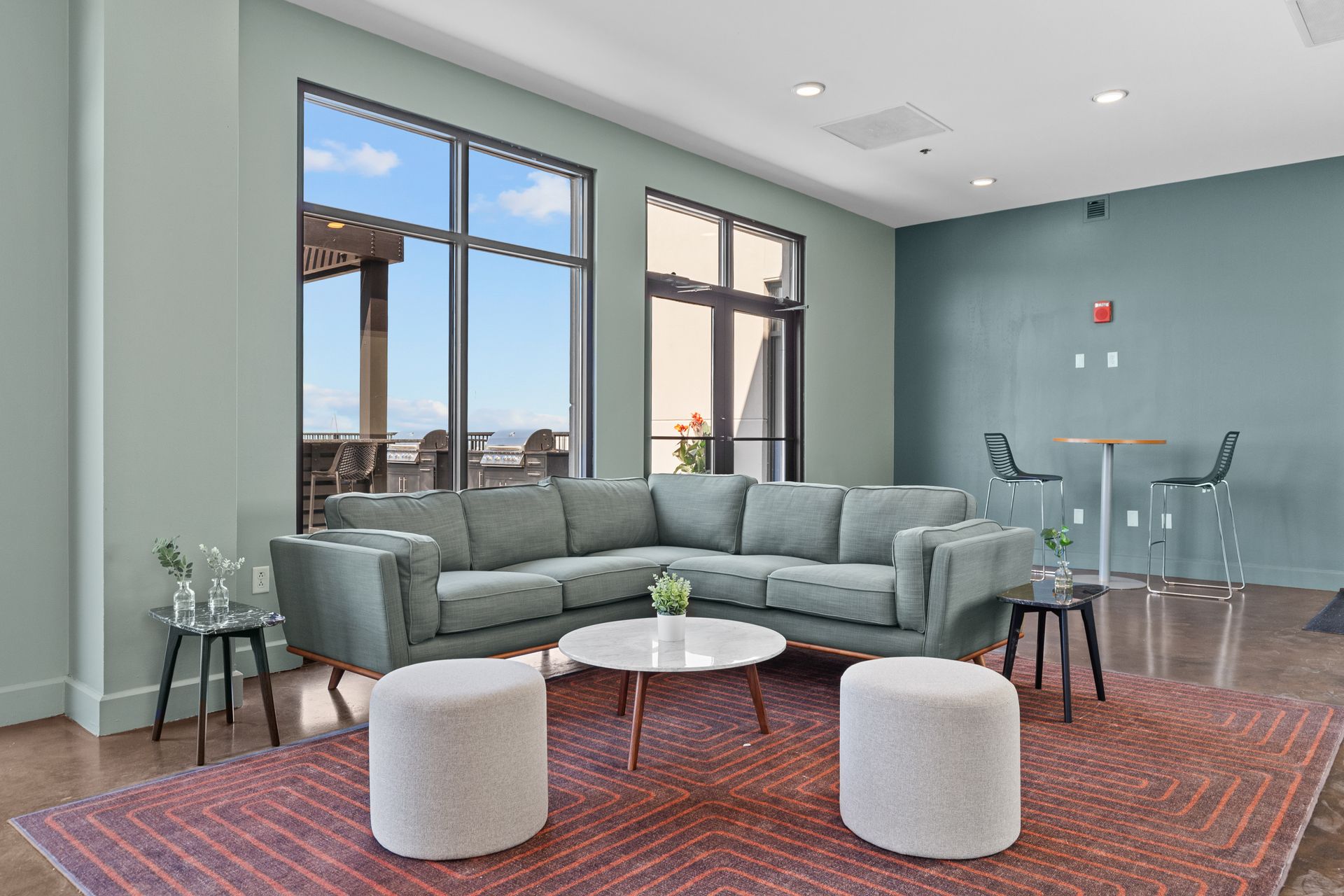 Modern lounge with gray sectional sofa, round coffee table, poufs, and large windows overlooking the city.