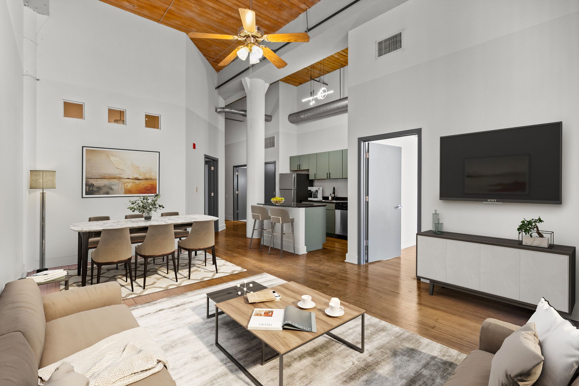 Bright open living room with beige sofa, dining table, kitchen, wall TV, and ceiling fan on wood floors