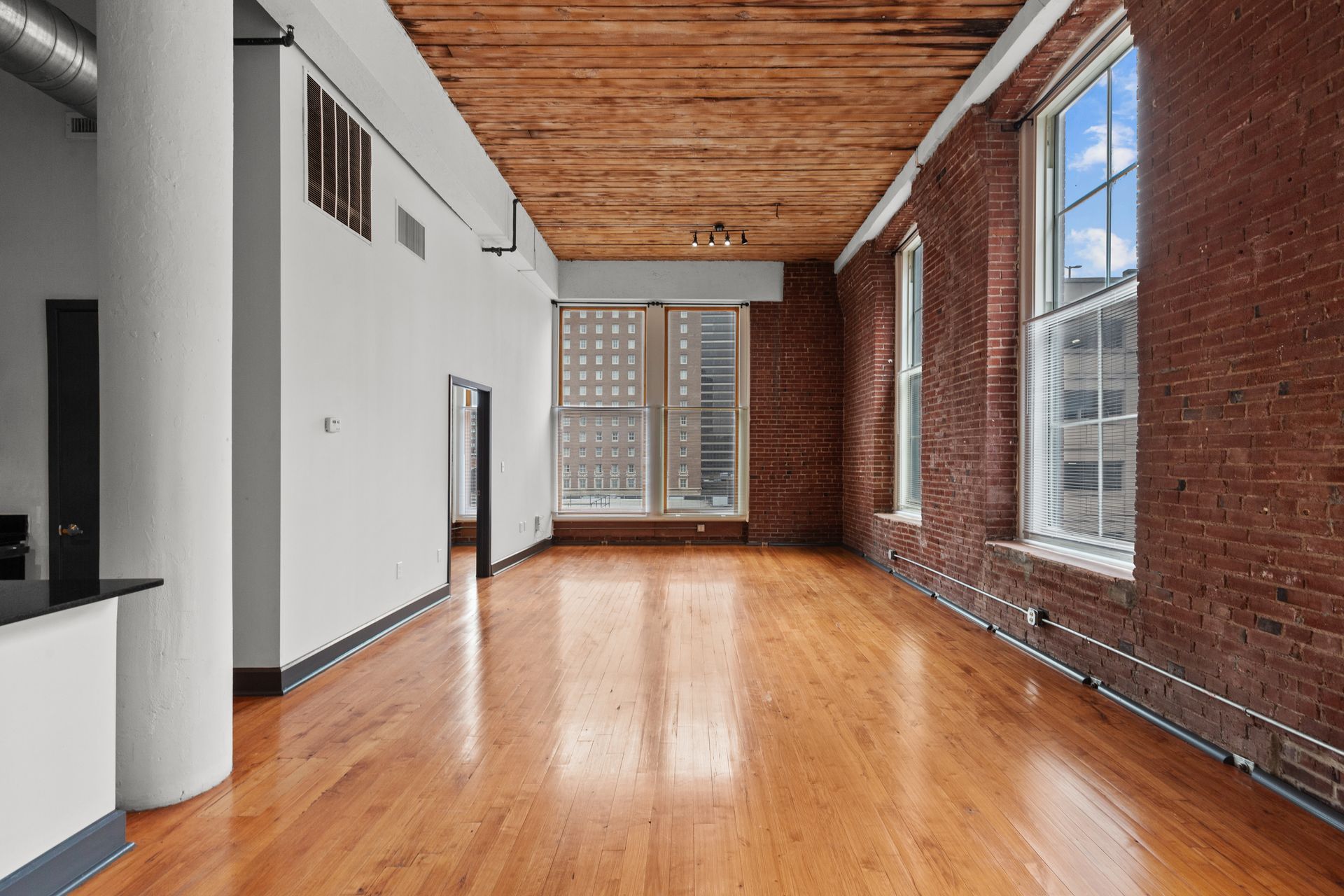 Empty loft with hardwood floors, exposed brick wall, and large windows letting in daylight.