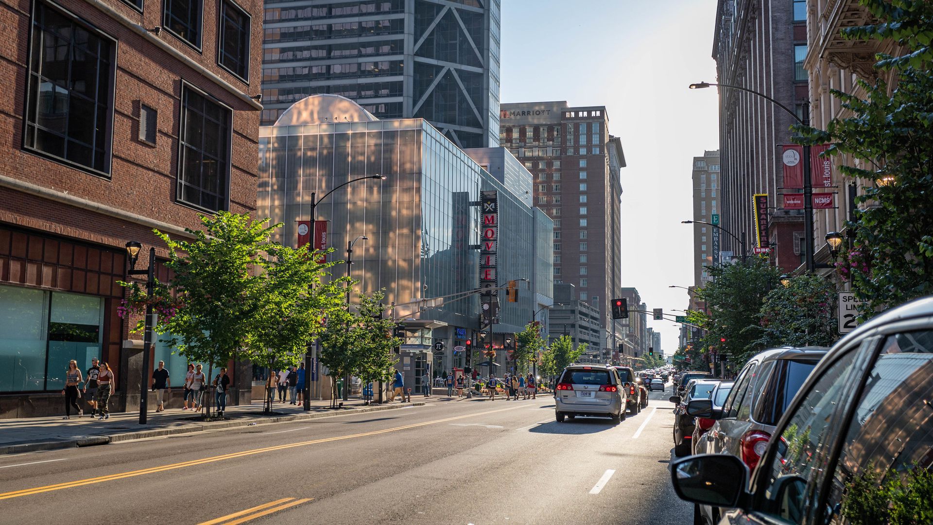 Sunlit city street with brick buildings, parked cars, and a few pedestrians downtown