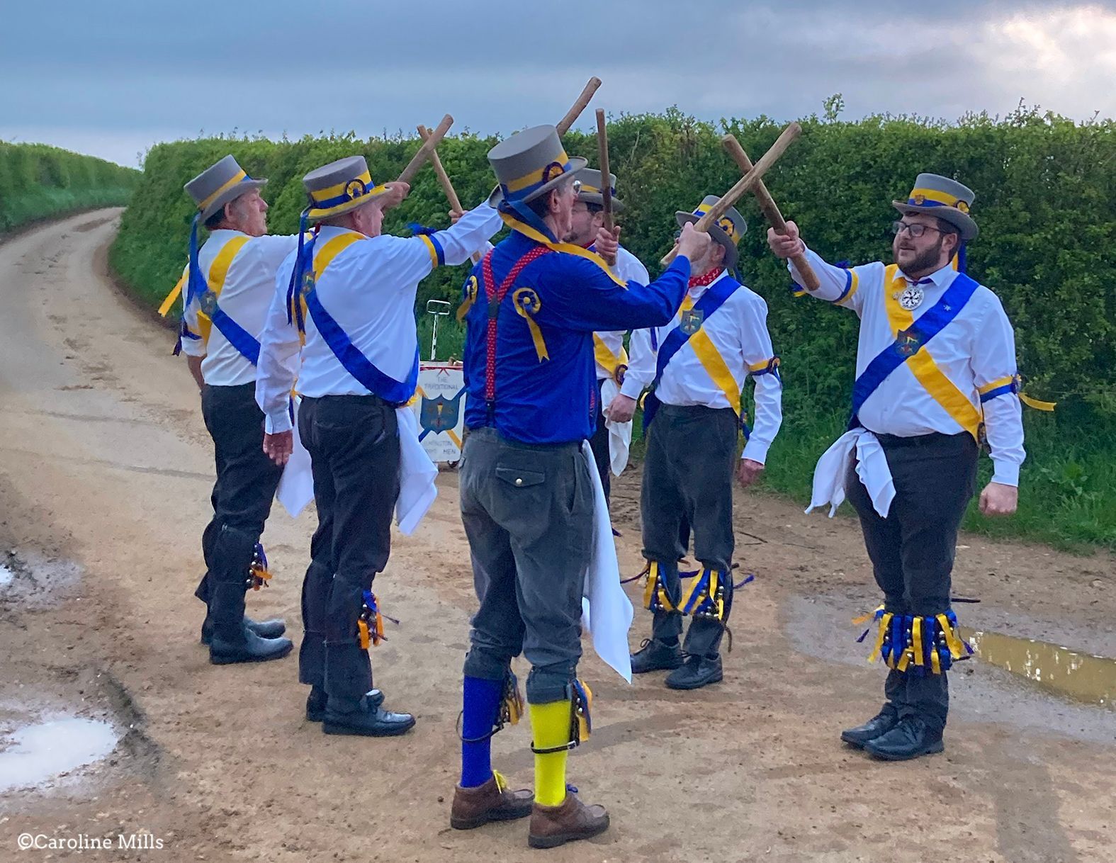 A group of morris men standing on a dirt road holding sticks