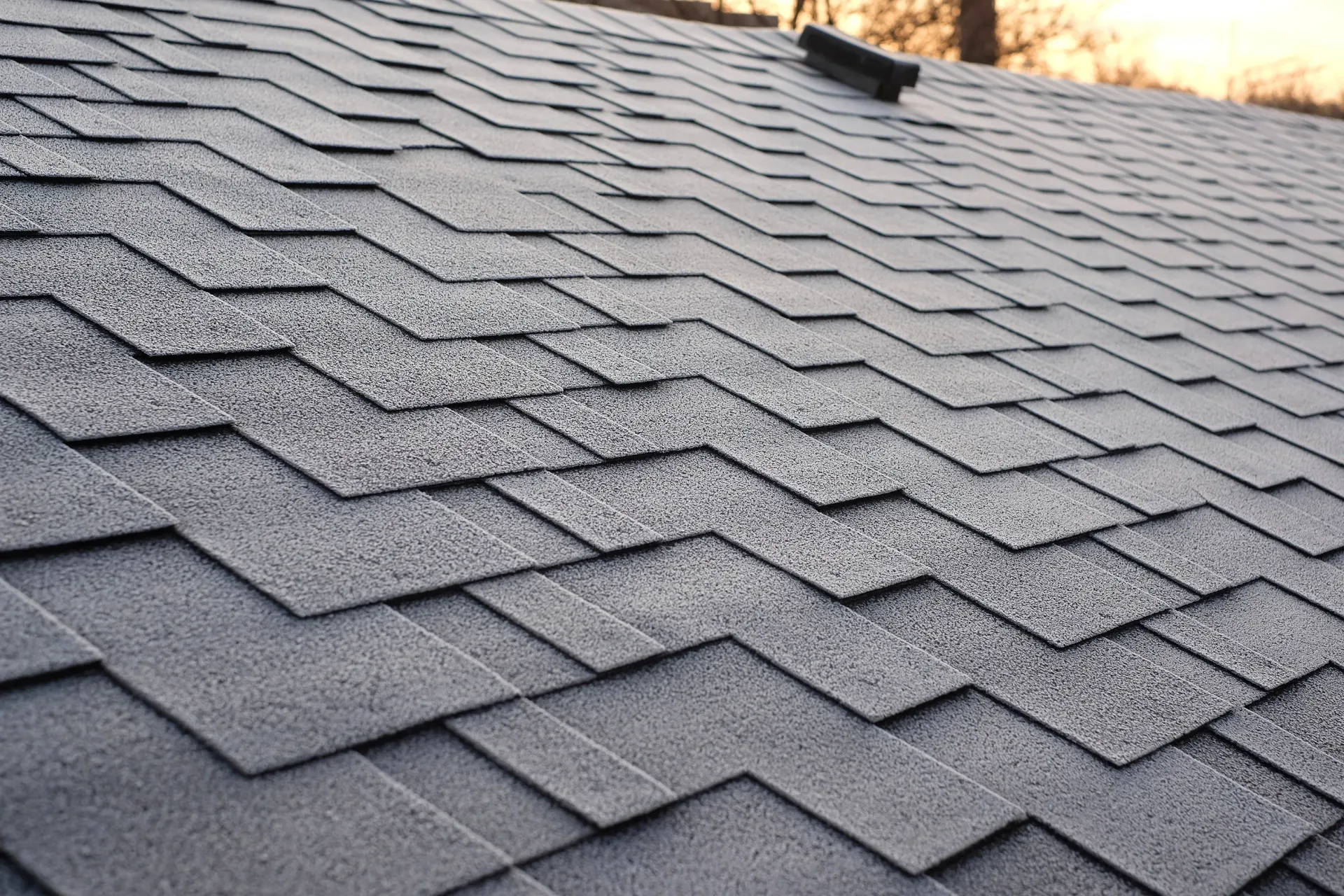 Close-up of gray asphalt roof shingles with a wavy pattern and a dark roof vent opening
