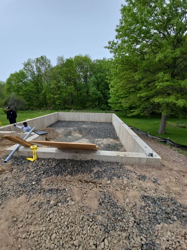 Concrete foundation site with gravel, wooden forms, tools, and trees in the background