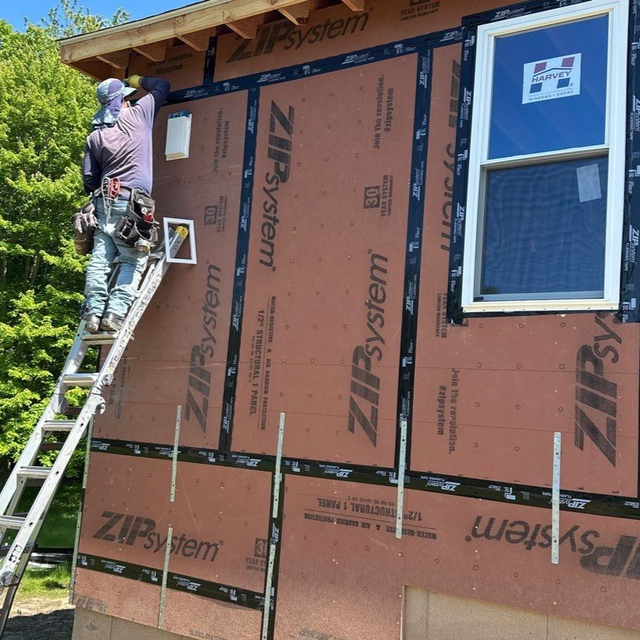 Worker on ladder installing ZIP System sheathing on a house exterior beside a window.
