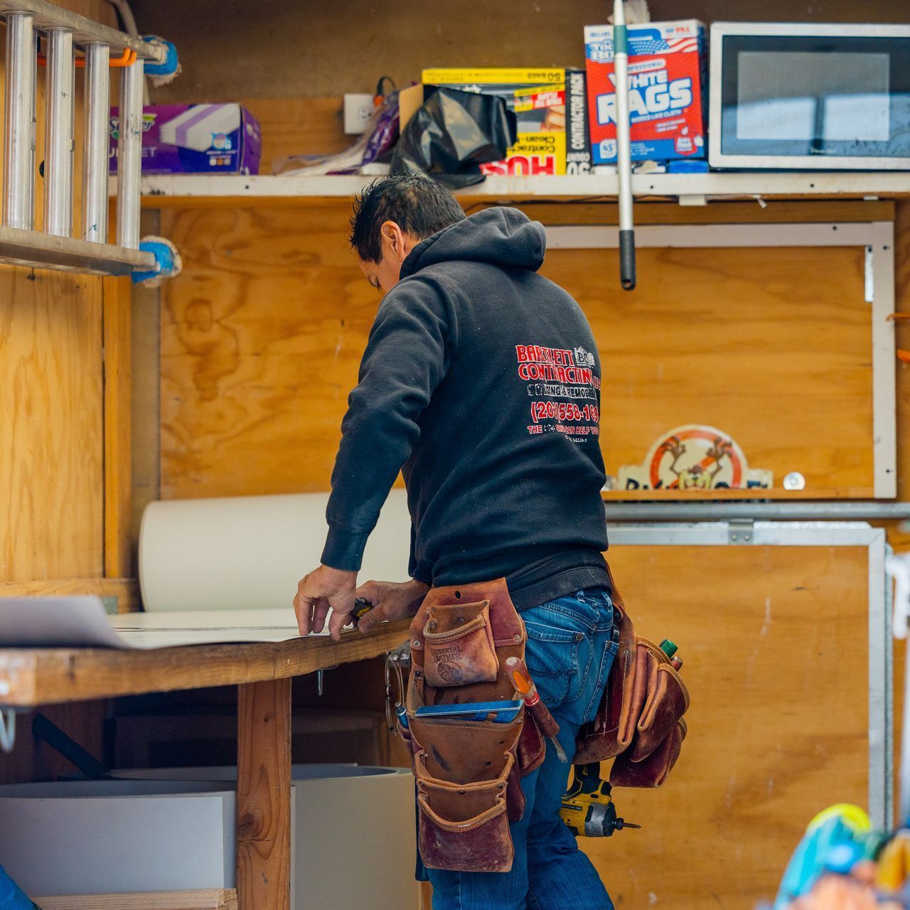 Worker in a black hoodie and tool belt measuring wood inside a workshop