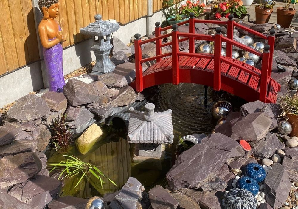 Red timber bridge over a pond edged with plum slate rocks in Bramhope