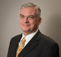 Man in suit and tie, smiling at the camera, against a gray background.