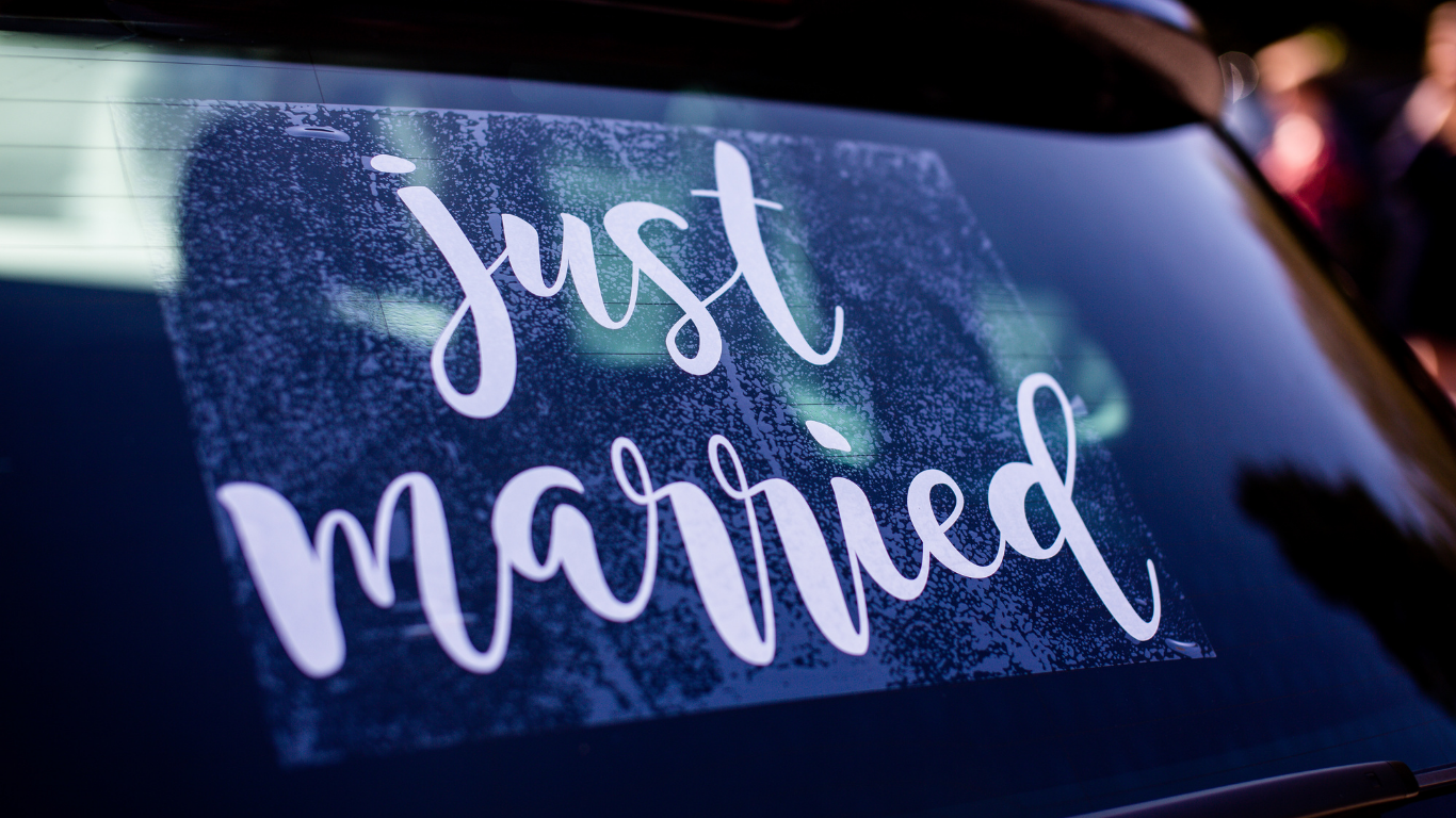 Bride in white strapless dress, holding bouquet, looking out car window, arm raised.