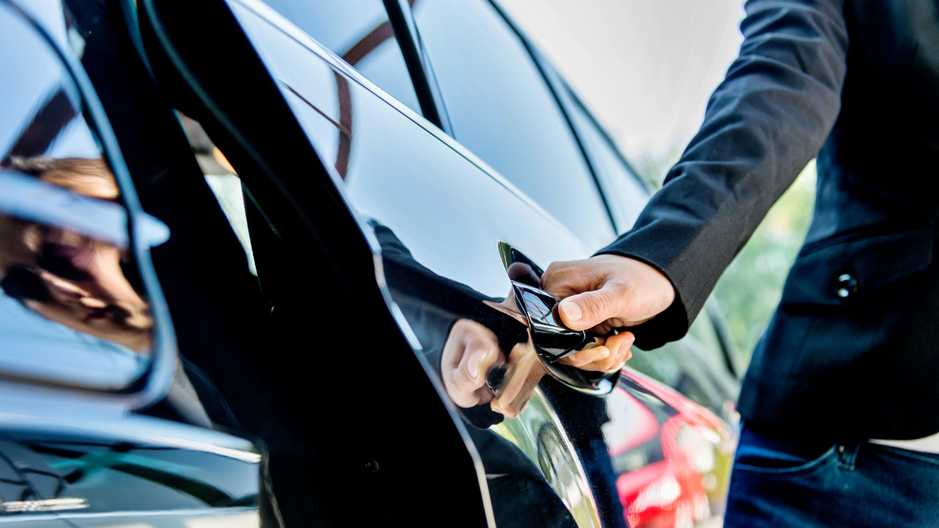 Couple taking selfie; woman in pink dress, man in suit with a car in the background.