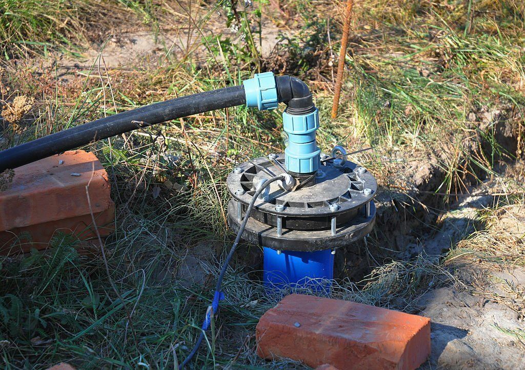 A blue plastic water well cap sits in the ground with a black pipe connected by blue fittings, surrounded by grass.