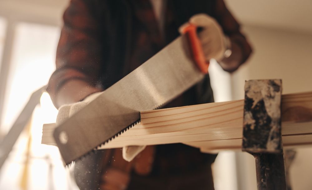 Person sawing wood with a hand saw, wearing gloves and a plaid shirt.