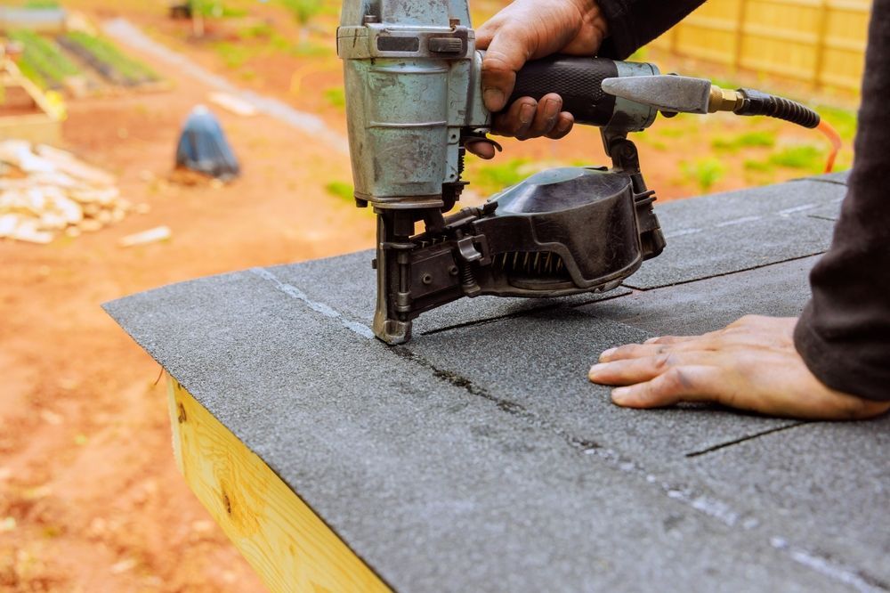Person uses a nail gun to install roofing shingles on an outdoor wooden structure.