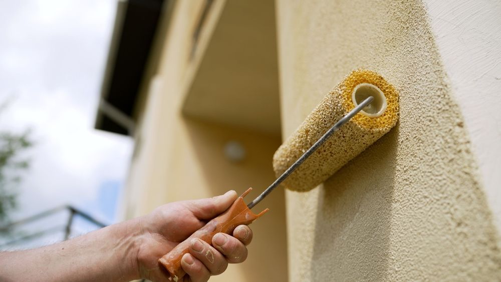 Hand using a paint roller on textured exterior wall, yellow paint.