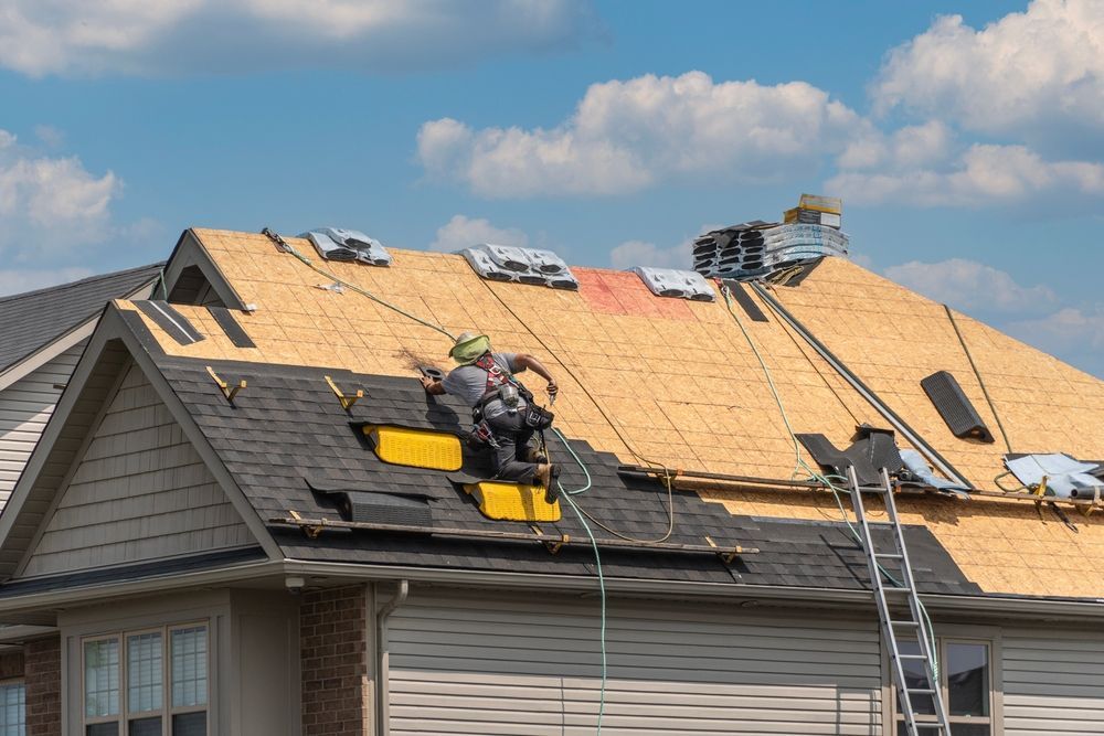 Roofers working on a house roof. One is secured by a harness, laying shingles. Ladder, blue sky, and materials are present.