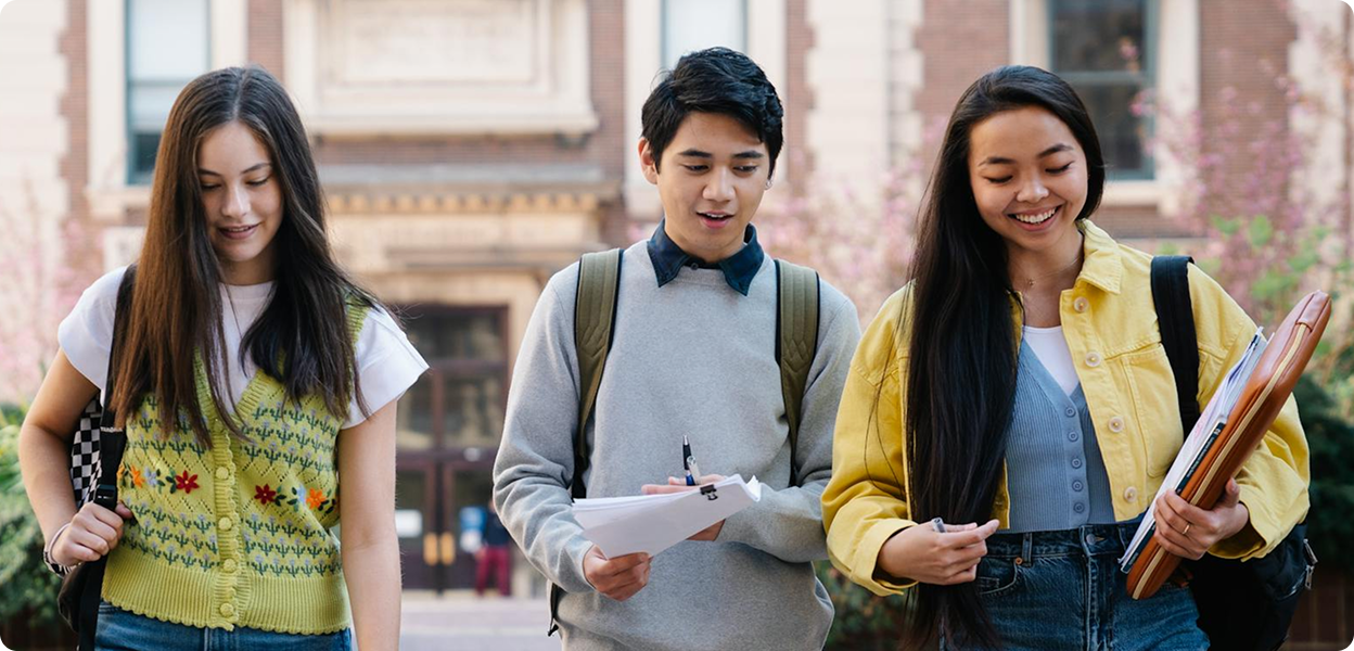 Three students walking down steps in front of a brick building; one carries papers, one a folder.