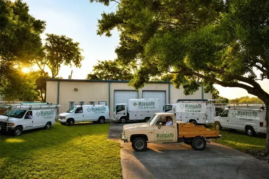 Fleet of white utility trucks parked on grass beside a beige building under large trees at sunset