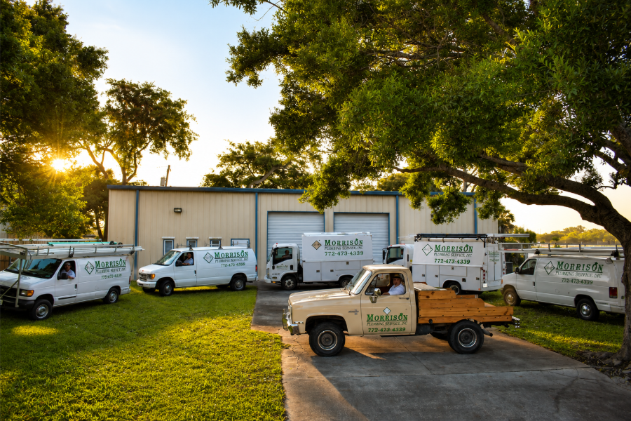 Fleet of white utility trucks parked on grass beside a beige building under large trees at sunset