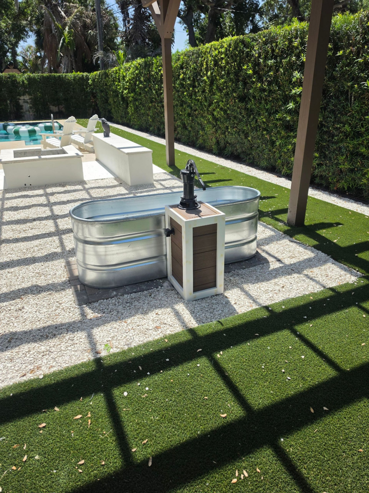 Outdoor patio with silver spa tubs, white lounge seating, and hedge-lined walls under bright sun.