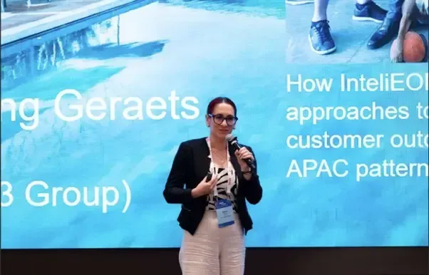 Woman speaking on stage in front of a blue presentation screen at a conference