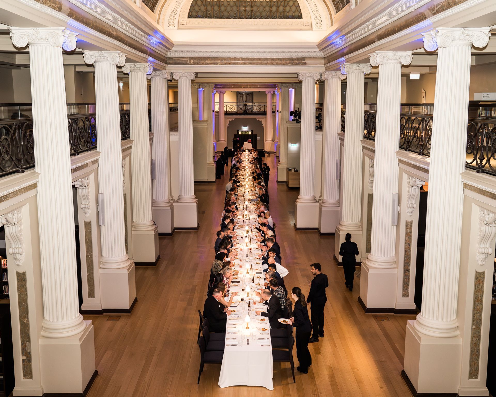 Elegant banquet hall with a long white-clothed table and guests beneath tall white columns and ornate ceiling.