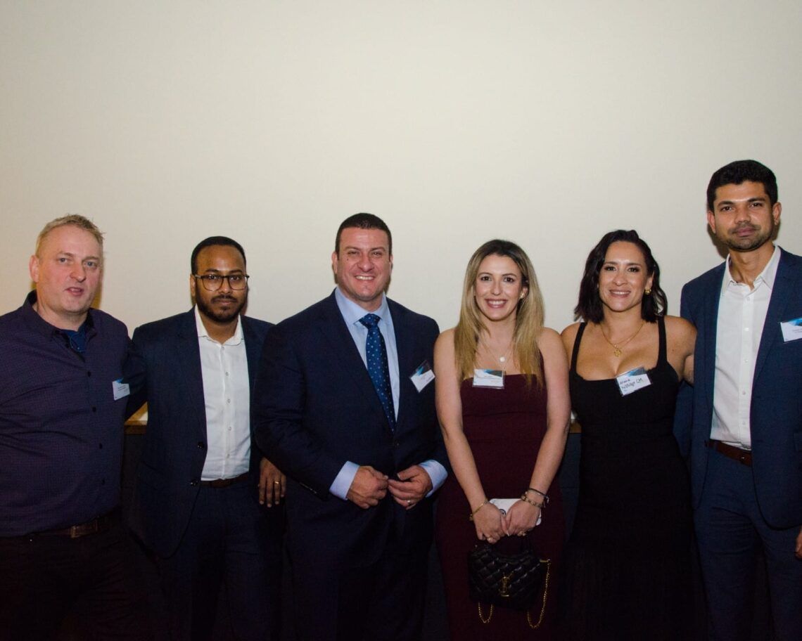 Six people dressed formally pose together at an event against a plain backdrop.