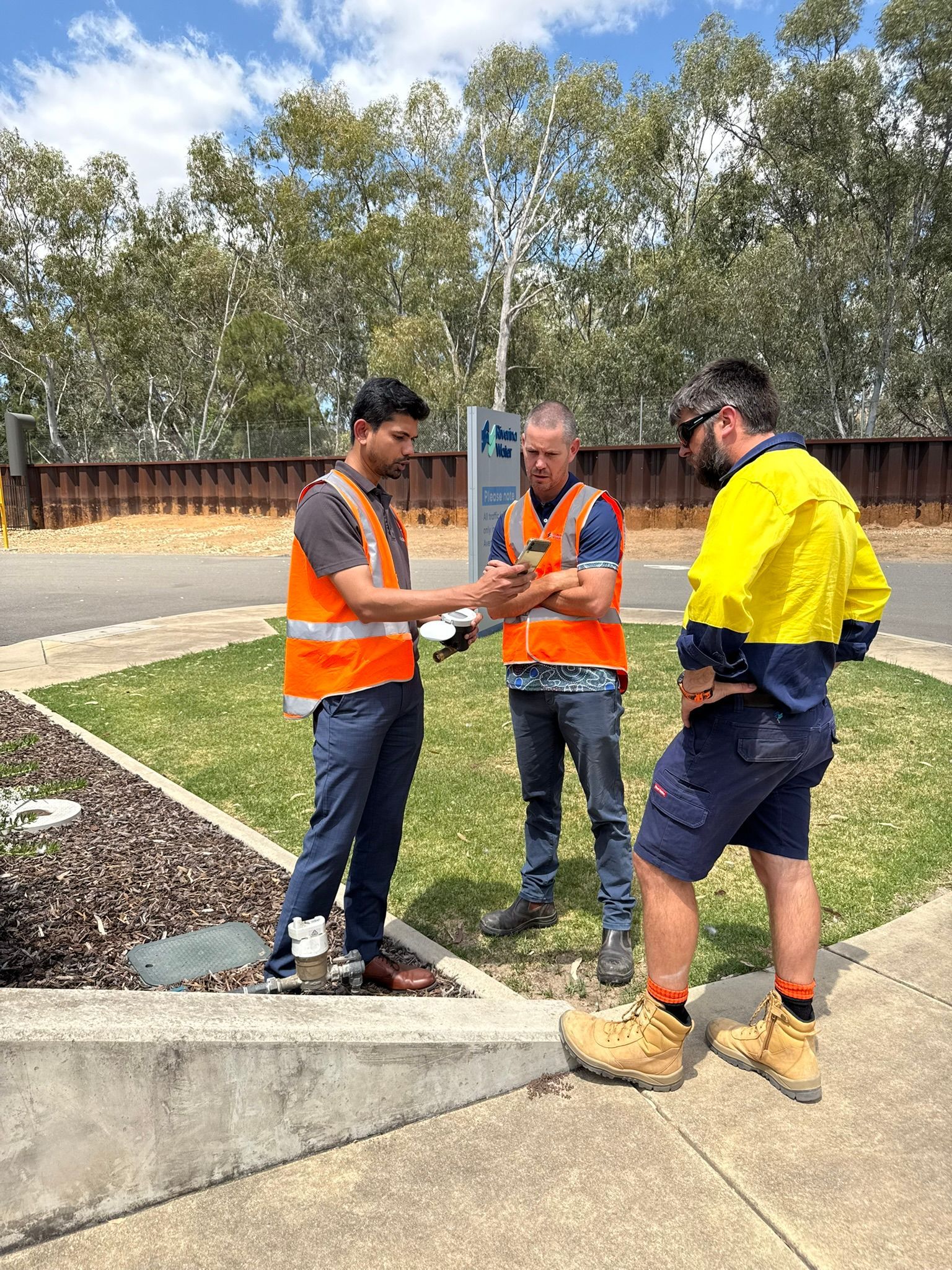Three workers in safety vests and boots discuss plans at a construction site.