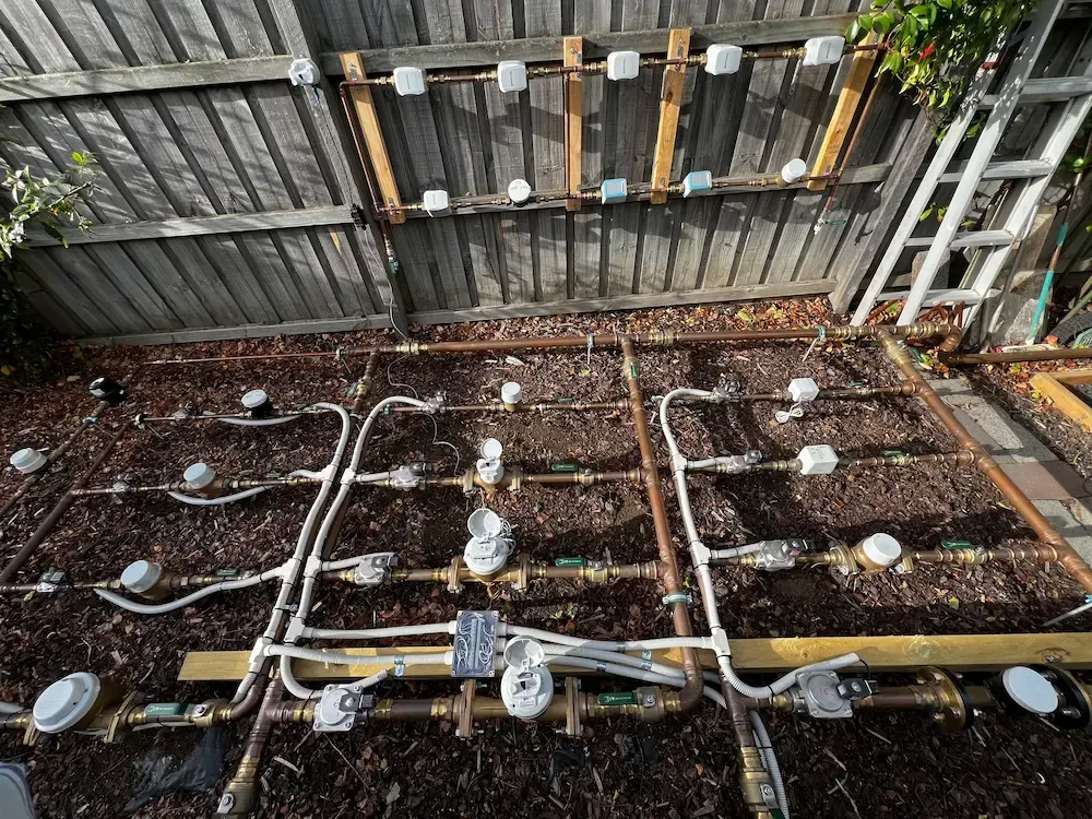 Rows of gas meters and pipes mounted on a beige wall outdoors.