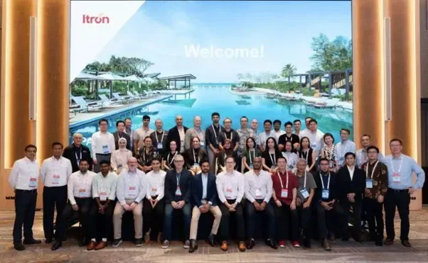 Large group posing in a conference hall before a tropical resort backdrop, with “Welcome!” displayed on screen