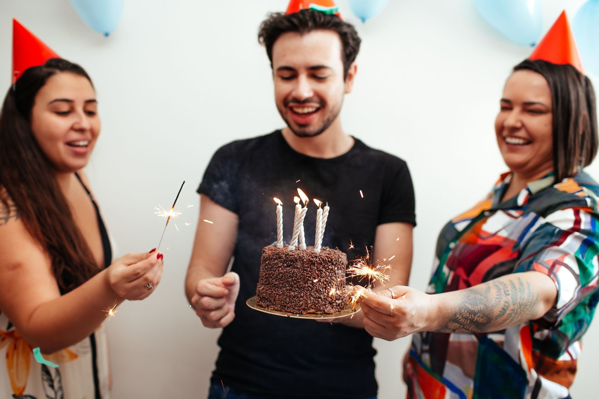 Three people in party hats smile around a chocolate birthday cake with lit sparklers.