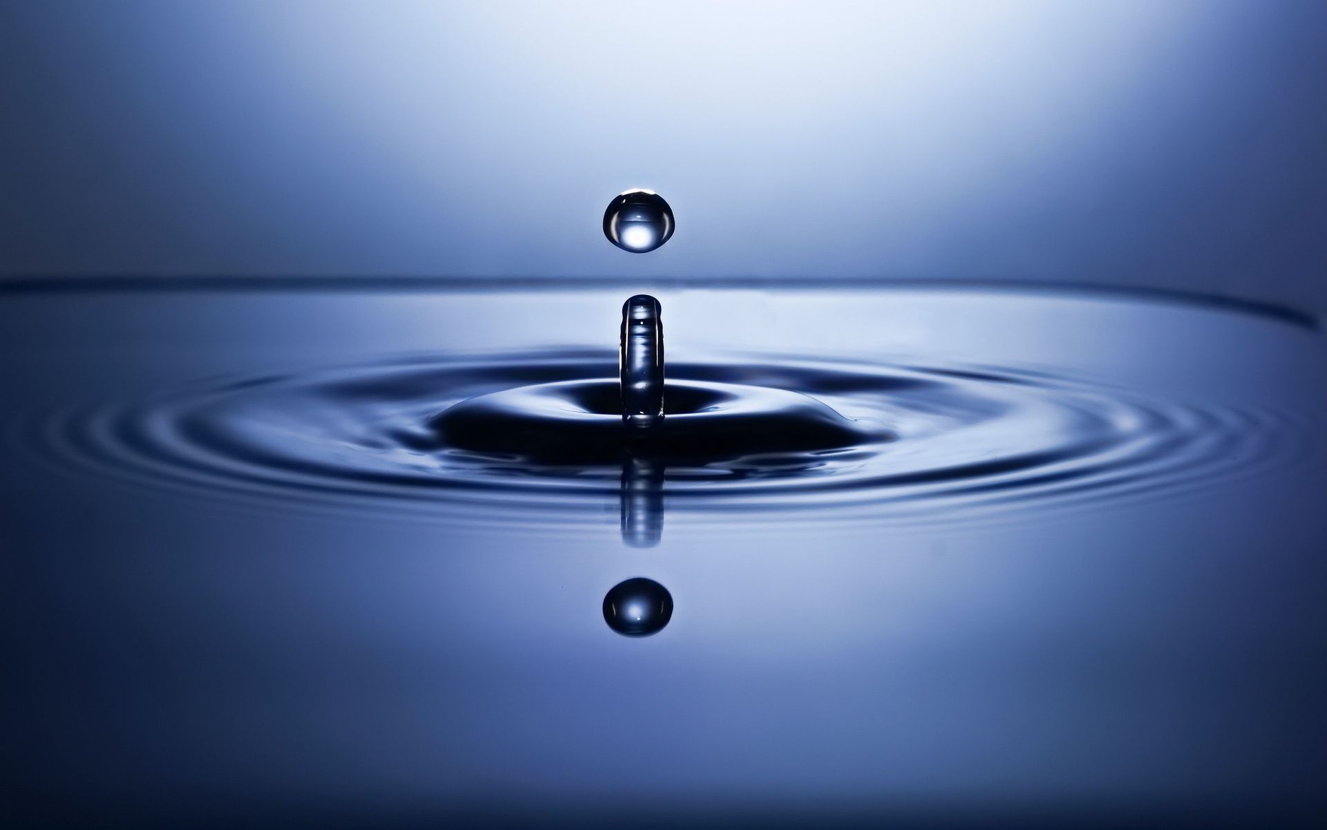 Water droplet splashing into a calm blue surface, creating ripples and a mirrored reflection.