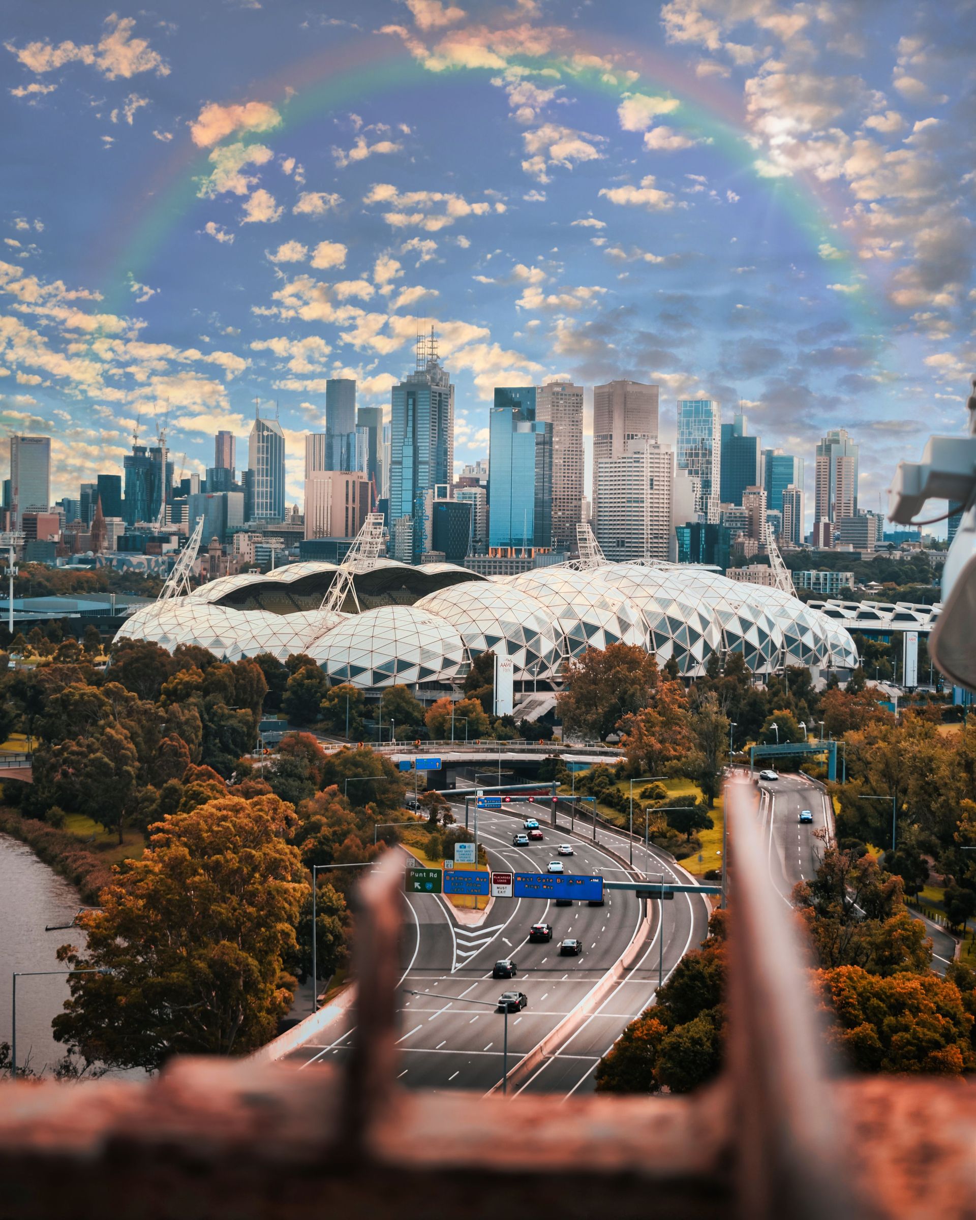 City skyline behind a white stadium and tree-lined roads under a colorful cloudy sky.