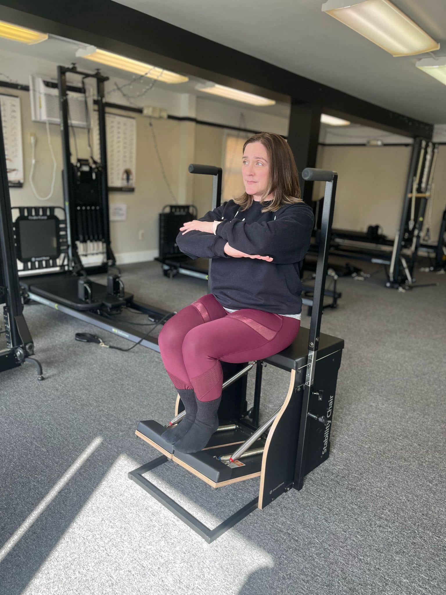 Woman seated on a Pilates chair, arms crossed, exercising. Maroon pants, black top, in a studio setting.