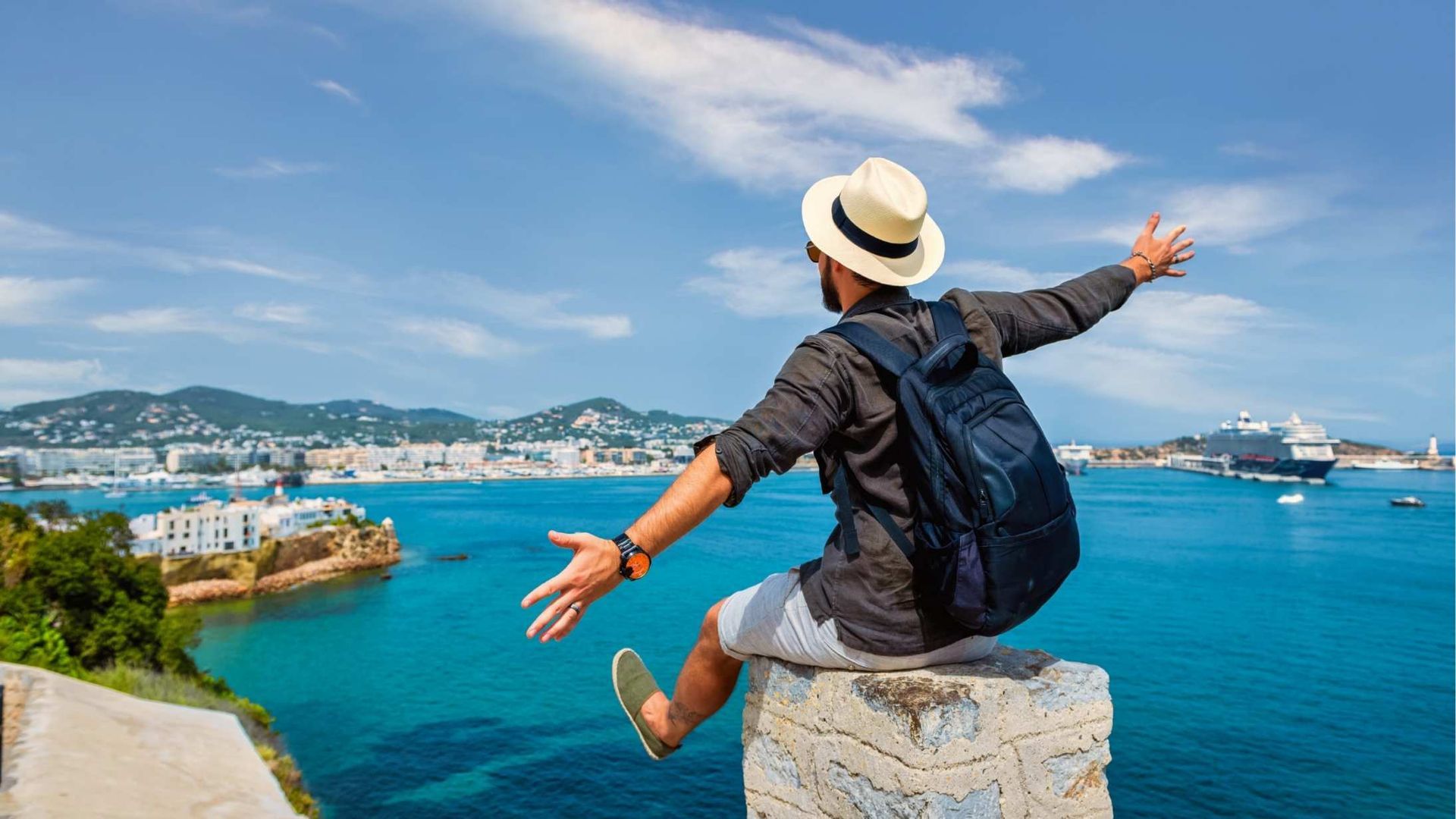 Man with arms outstretched, wearing a hat, overlooking turquoise water and a coastal city under a blue sky.