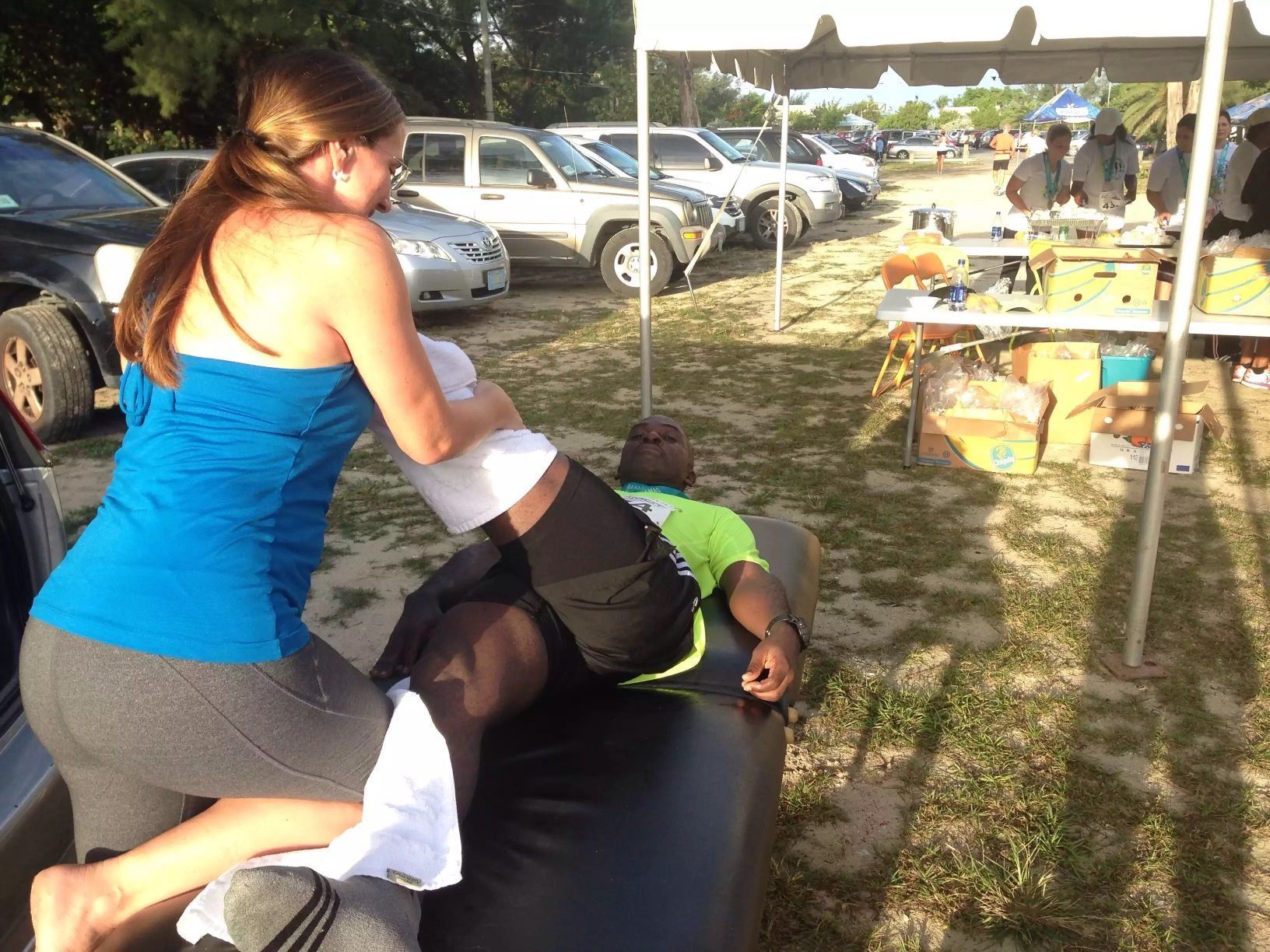 Woman giving a leg massage to a man outdoors, cars and tent in background.