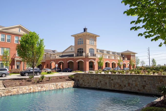 A multi-story brick commercial building with a central tower overlooks a tranquil pond with a stone retaining wall.