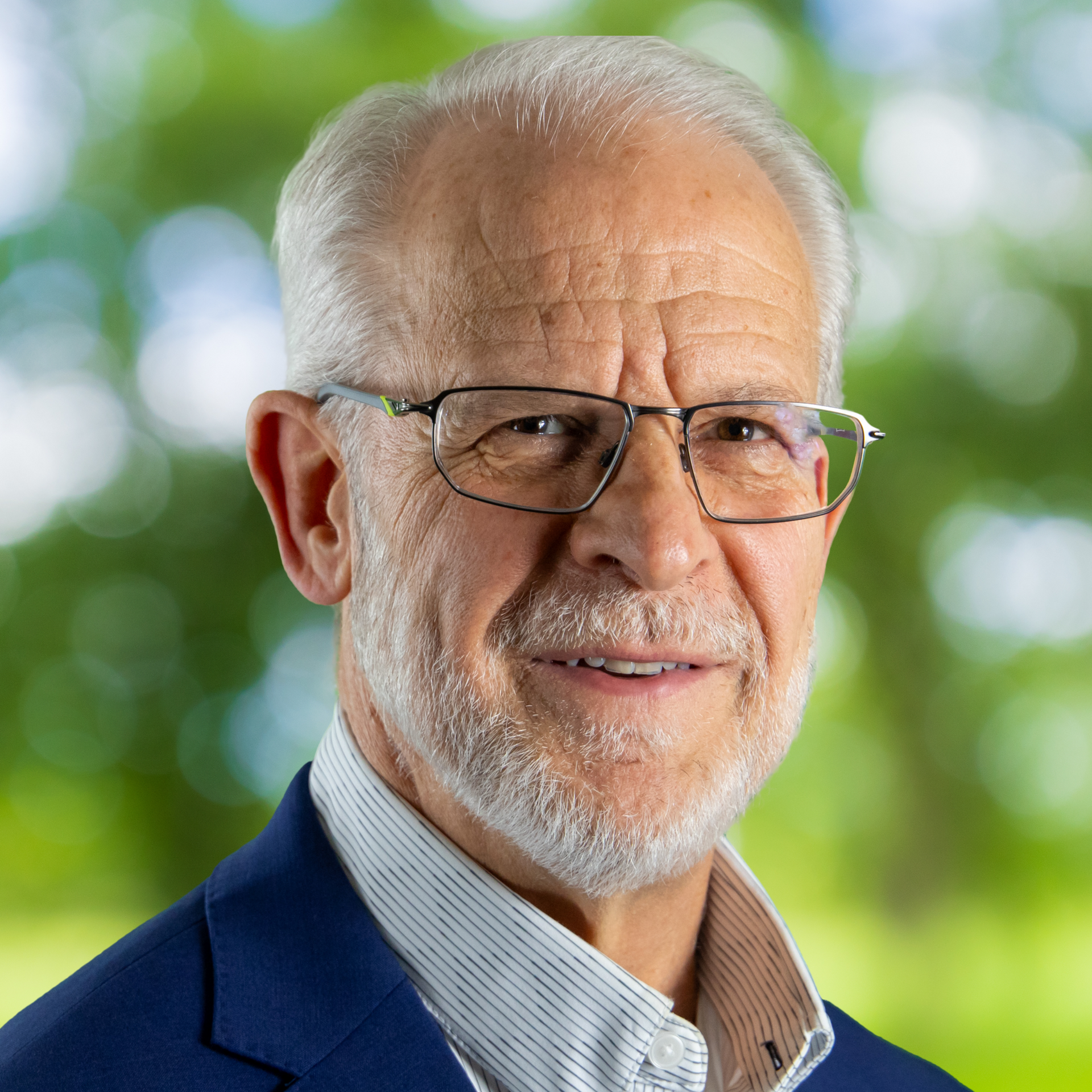 A man with a white beard and glasses wearing a patterned blue and white shirt stands in front of a green leafy backdrop.