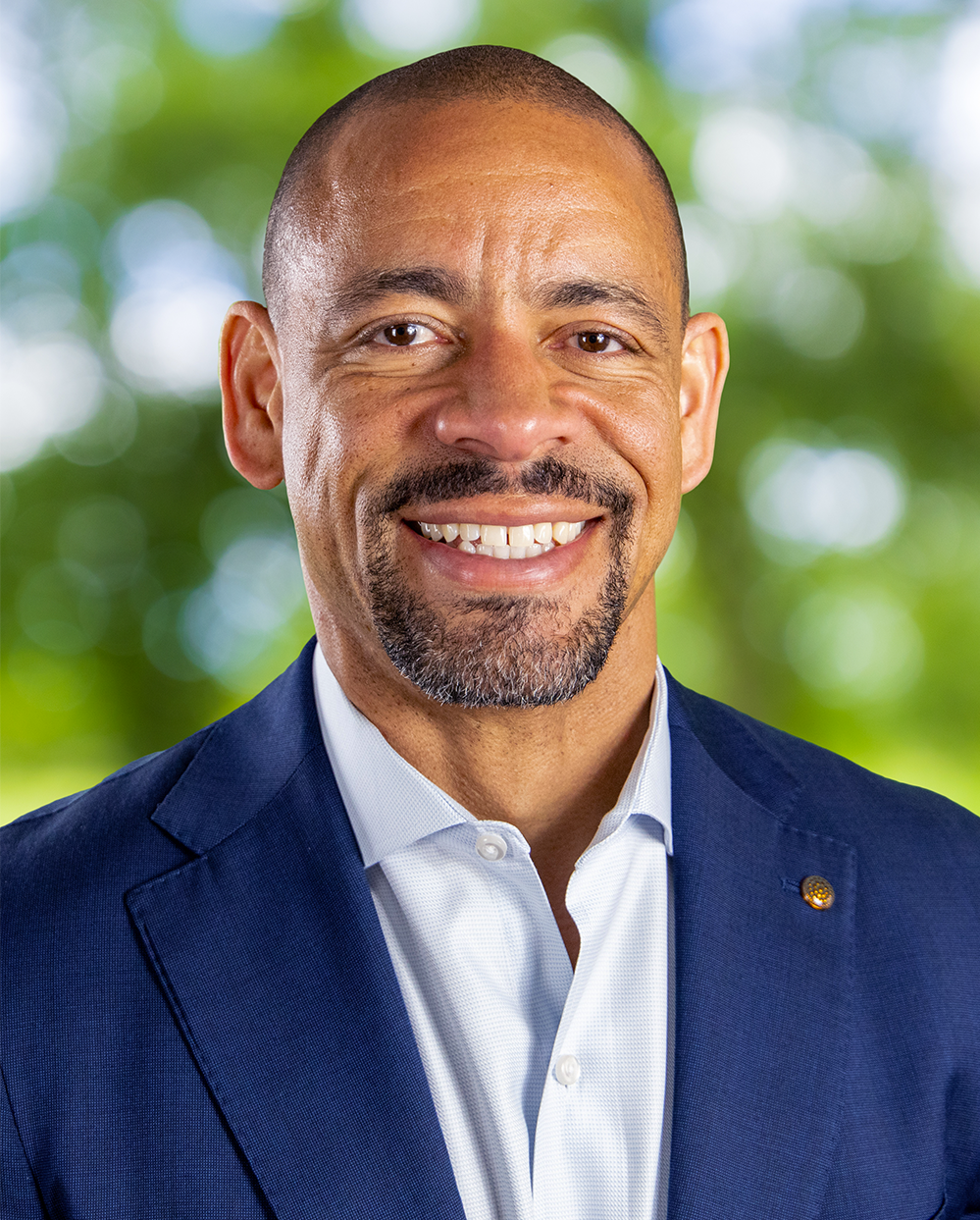 Smiling man wearing a blue blazer and light purple dress shirt with a pocket square, against a green leafy background.
