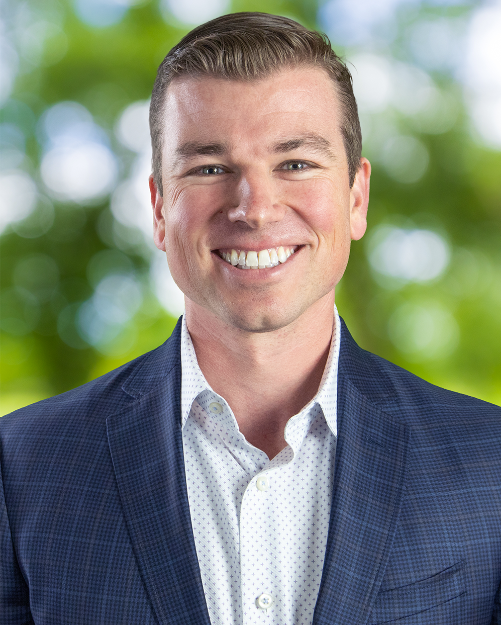 A person wearing a blue textured blazer and white collared shirt smiles against a soft-focus green leafy background.