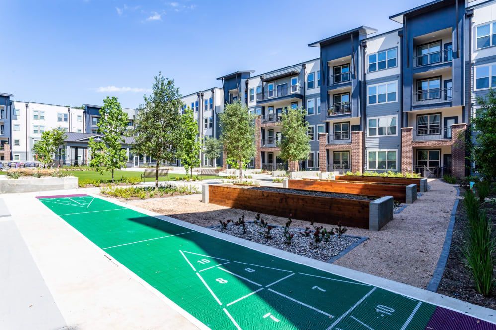 A green shuffleboard court sits in an outdoor courtyard between modern, multi-story apartment buildings.